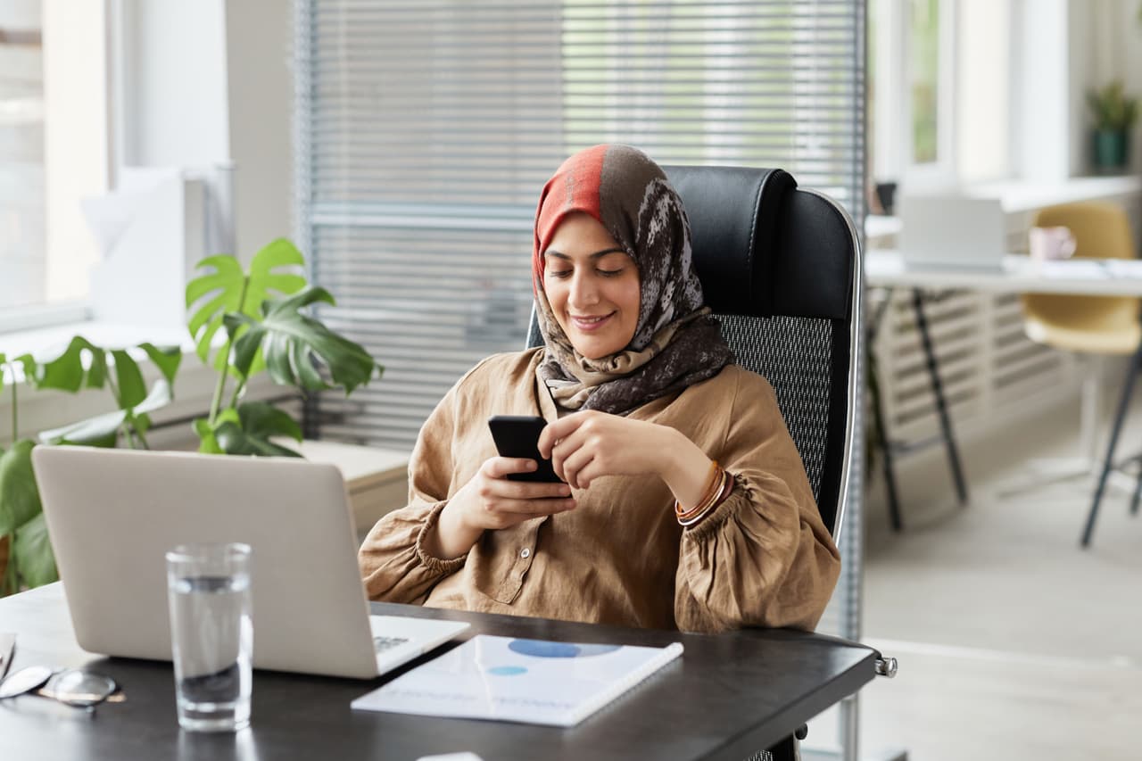 امرأة ترتدي حجابًا تبتسم وهي تنظر إلى هاتفها على مكتبها